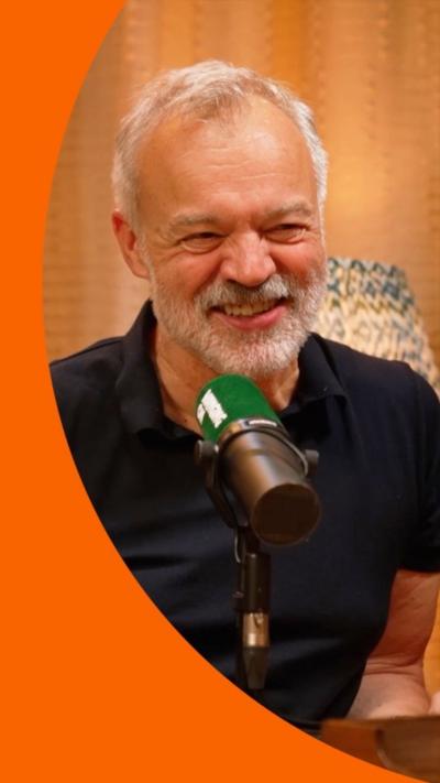 Graham Norton has white hair, a moustache and beard. He is wearing a dark-colour T-shirt and sits behind a microphone with a green muffler.