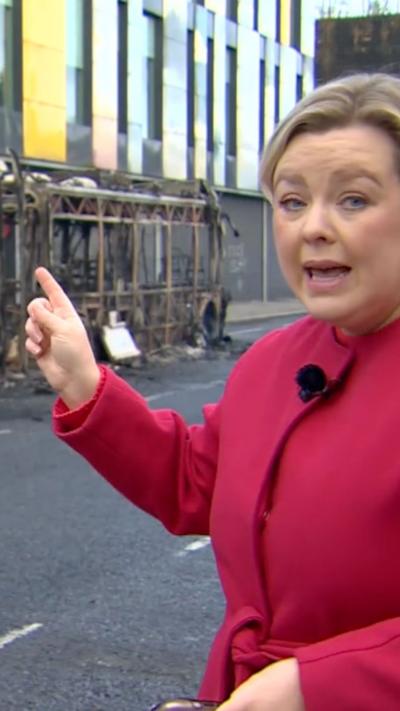 BBC correspondent Sara Girvin wearing pink coat and pointing at burned down bus