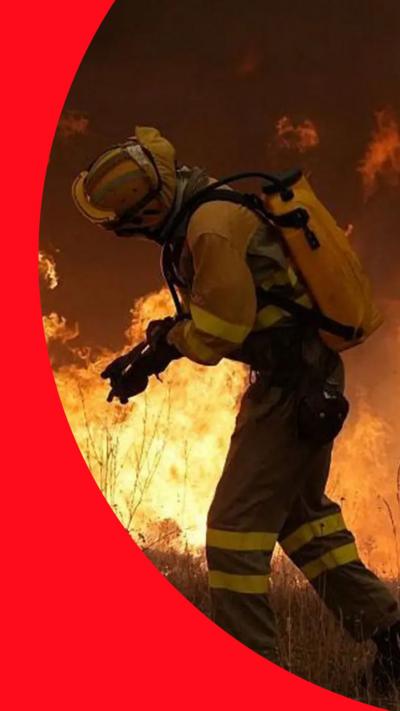 Firefighter walks in front of a large wildfire as flames spread through dry grass.