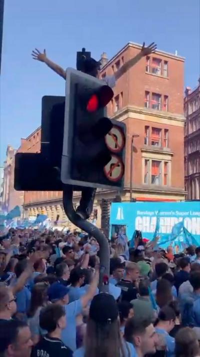 A crowd of people in blue are stood on the street, a man can be seen stood on top of the traffic light which is on red, with both of his arms in the air 