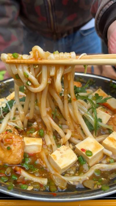 A pan of hot udon with cubes of tofu, with chopsticks lifting some noodles. 