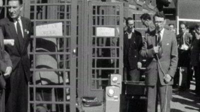 A reporter standing in front of telephone boxes.