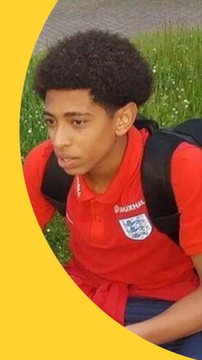 A boy with curly hair wearing a red football shirt sits on grass
