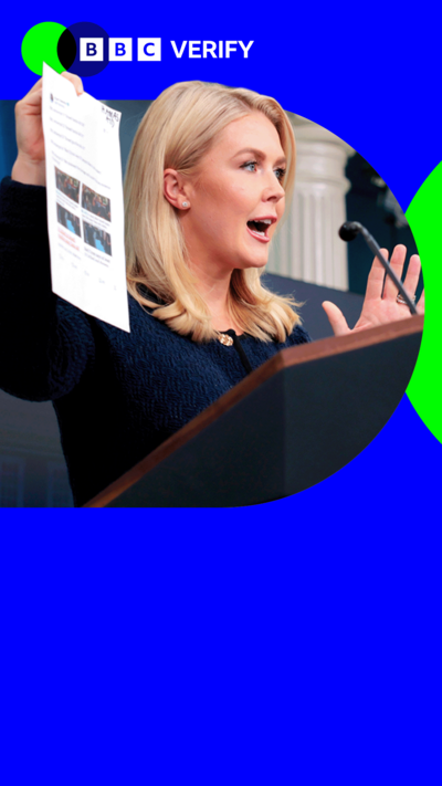 Karoline Leavitt holds up a print out of the X social media site showing BBC News articles. She is standing behind a lectern. 