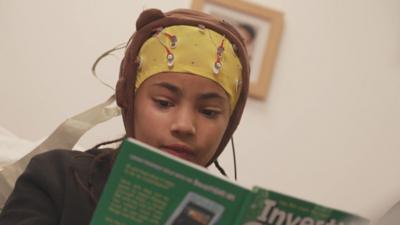 A girl wears a cap with wires coming out of it on her head. She is reading a book.
