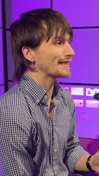 A person with medium-length hair wearing a checkered button-up shirt sits in a studio setting with purple lighting, speaking into a nearby microphone.