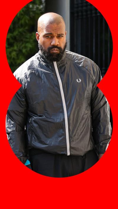 Kanye, or Ye, West has a shaved head and a black bear. He is a wear black trousers, and a black windbreaker with a white zip.