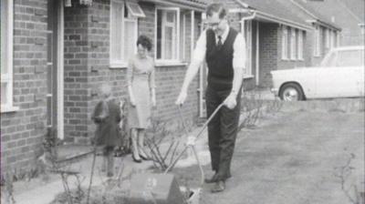 Philip cutting the grass in front of a house while his wife and child look on