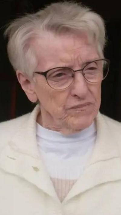 A woman with grey hair, wearing a cream jacket and white tshirt