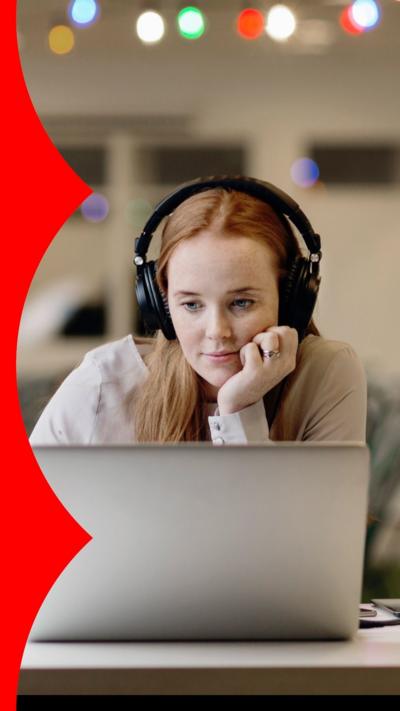 A woman in an office wearing headphones looks at her laptop. She has long red hair