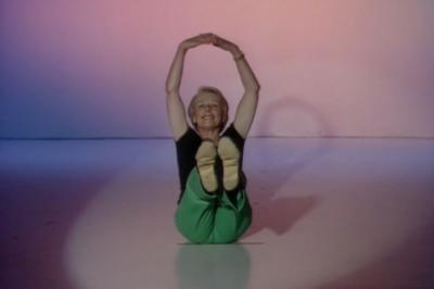 Eileen Fowler sitting on the floor doing aerobics, with her hands above her head and her legs in the air in front of her.