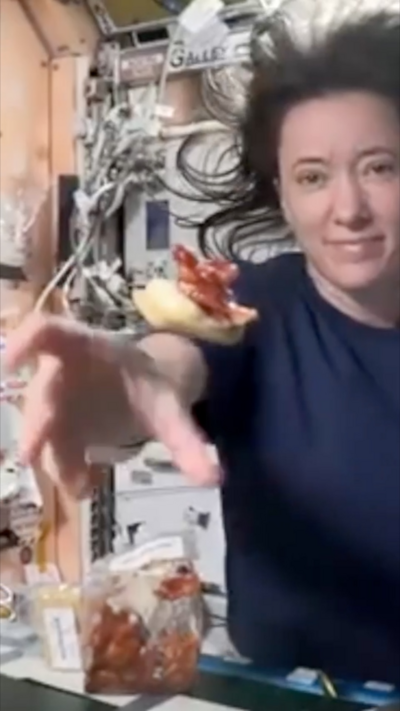 A woman floats in zero gravity, reaching for a bun and some meat floating in space in front of her. She wears a blue T-shirt