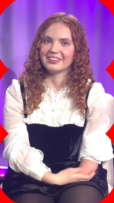 A woman, Bridgerton actor Gracie McGonigal, smiles. She has long curly red hair and wears a white shirt with a black dress over the top.