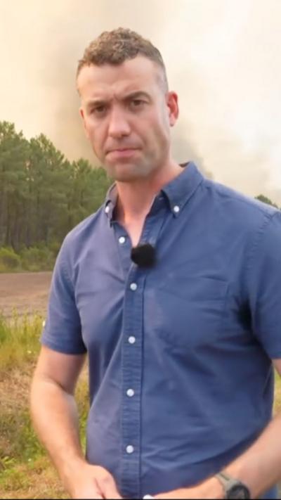 Gwyn stands in front of a forest from which black smoke rises in the background