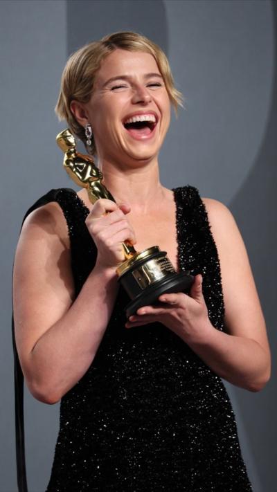 Jessie Buckley wears a sparkly black dress and has blonde hair. She laughs as she holds up her Oscar