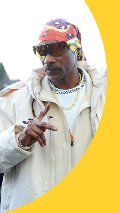 Snoop Dogg wears a multicoloured bandana, shades and a cream coat 