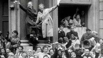 Crowds of people stand in front of a building. Two women stand on a pillar