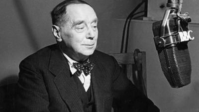 Black and white image of HG Wells is 3 piece suit and bow tie, sat in front of a microphone with 'BBC' logo on it.