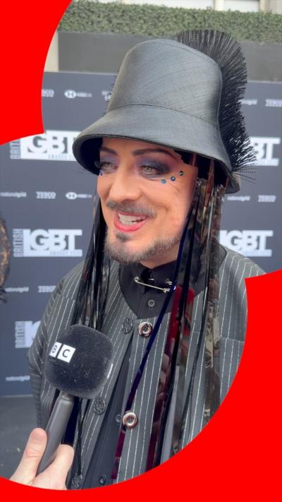 Boy George wears a black cape and hat with heavy dark makeup