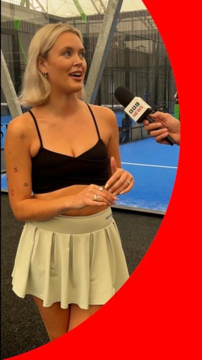 A woman in a tennis skirt and black strap top, in a padel stadium, talks into a BBC News microphone.