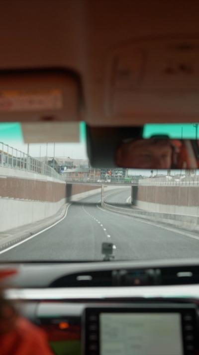 The view from inside a vehicle as it travels down a newly built underpass, with the road curving gently ahead and concrete walls on both sides. The dashboard and steering wheel are visible in the foreground, along with the reflection of the driver’s face in the rear‑view mirror.