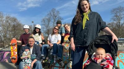 A group of mums sit together at a skate park. One mum has a baby in a pram. 