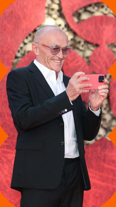 Danny Boyle, wearing a suit and glasses, stands smiling with a smart phone in his hands