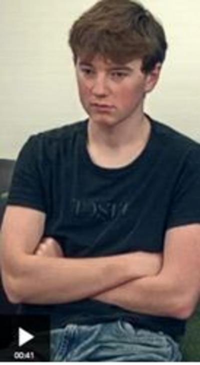 A teenage boy sits with his arms folded looking down. He has brown hair and eyes, and is wearing a black t-shirt and blue jeans.