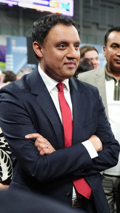 Anas Sarwar photographed at a press conference, wearing a black suit, white shirt and red tie. The photograph is a close-up shot of his head and neck.
