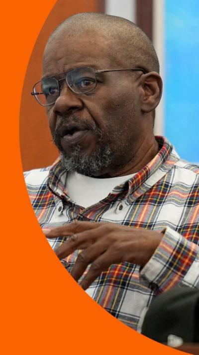 A man wearing a checkered shirt and glasses testifies in court.