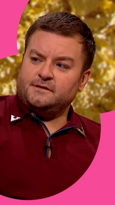 Alex Brooker in front of a gold background