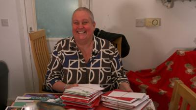 A woman with a buzzcut smiles at the camera. The table in front of her has loads of cards and letters. She has a green, blue and white patterned shirt on.