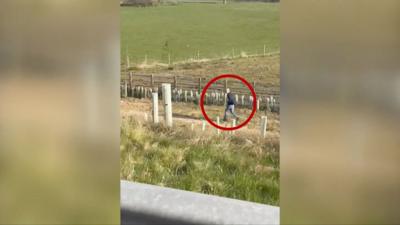 A screengrab of a mobile phone video showing a man in a field next to a road.