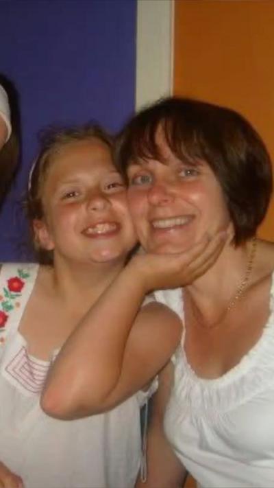 A young girl, wearing a white blouse and with her hair tied back, is smiling and has her hand cupping an older woman's chin. The older woman has a bob haircut, with reddish hair, also wearing a white top.