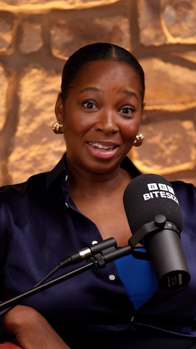 Jamelia looking at the camera with a surprised look on her face. Microphone in front of her, blue shirt and top. She's sitting a podcast studio with a brickwall effect behind her.
