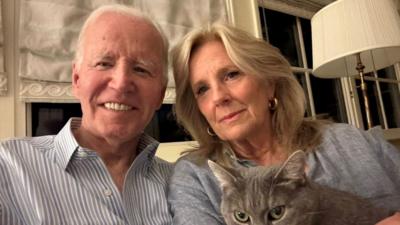 Joe Biden and Jill Biden pose for a selfie photo. They are sat down in front of a curtained window, Jill is holding a grey cat.