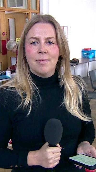A blonde woman holding a microphone and a phone. She is smiling at the camera and wearing a black top.