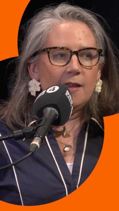 Viv Groskop has long grey hair and is wearing glasses with brown patterned frames and earrings that look like white flowers. She is wearing a navy v-neck top with white piping details.