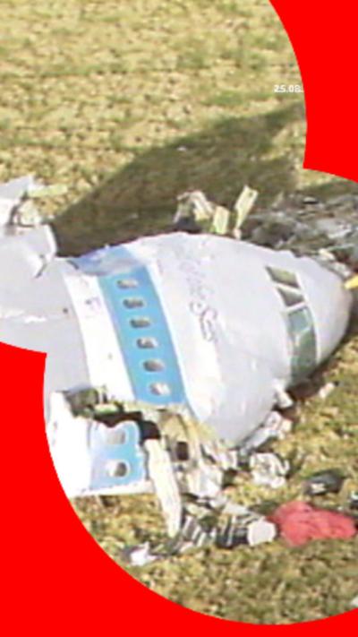 Archival image - a white plane with a light blue stripe, crashed in a field. Only the partial front end can be seen with debris strewn around.