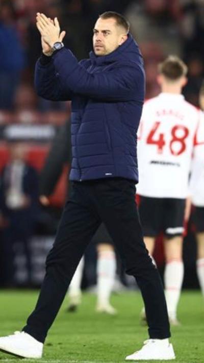 Tonda Eckert walks across a football pitch with his hands raised, clapping. He has short brown hair and wears a navy blue hooded puffer jacket and black jeans, with white trainers.