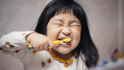 Young girl brushing teeth with yellow toothbrush, scrunching up her eyes in happiness.
