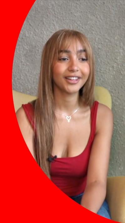 PinkPantheress has long brown hair and is wearing a red top and blue jeans.