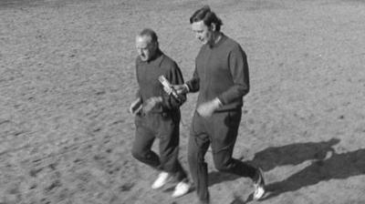 Two men wearing tracksuits jog across the grass in the park.