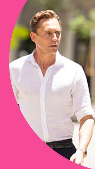 Tom Hiddleston walks in the sunshine. He wears a white shirt and black trousers