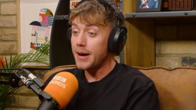 A man, Roman Kemp, wears a black top and sits on a sofa in front of a Sounds branded microphone
