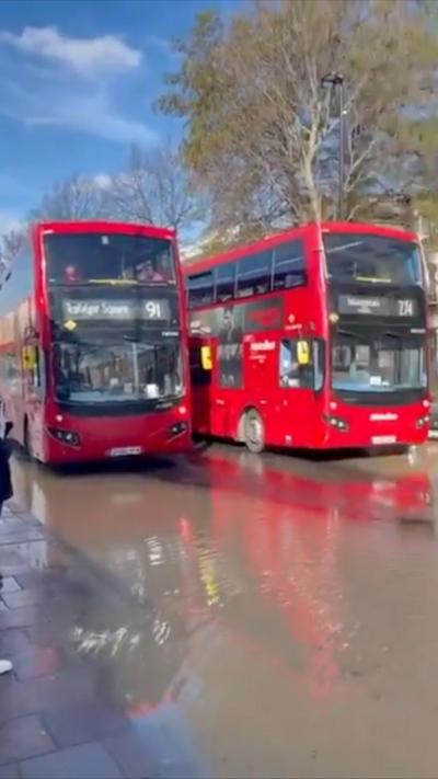 Two red double-decker buses sit above a thin layer of water, which has flooded the entire road.