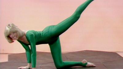 Diana Moran ('The Green Goddess') with her palms on a mat on the floor, lifting her left foot up back behind to point to the sky, whilst her right leg is laid on the floor.