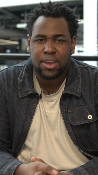 A man in a dark denim jacket and a beige t shirt is sat down looking straight at the camera. He has spiky afro hair and there is a small mic poking out of his t-shirt. In the background there are blurry tv screens with various different output from the BBC. There is also a glass wall roughly a waist height and two coloured squares, one blue and one green.