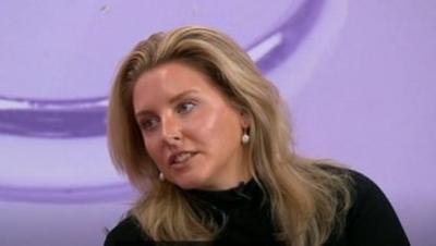 Emma, who has blonde hair and is wearing a black top, is sitting in front of a purple screen