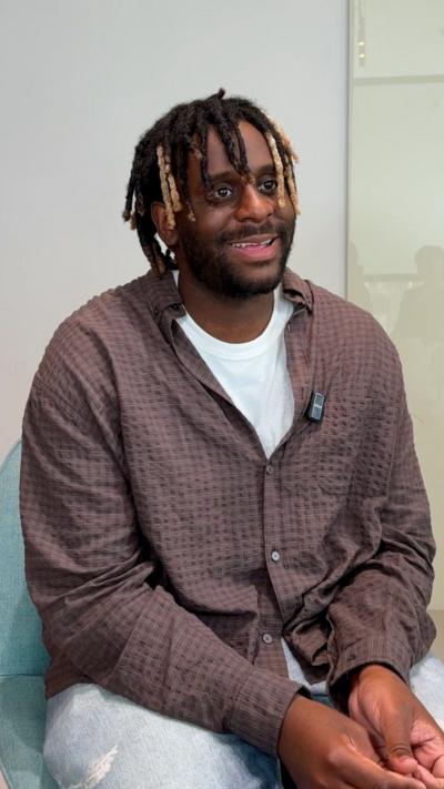 Myles Smith smiling. He is wearing a brown shirt over a white T-Shirt. 